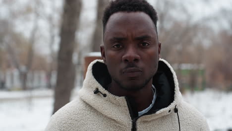 portrait of a young man with a bold look, wearing a winter jacket, standing outdoors with a blurred background featuring indistinct trees and urban structures