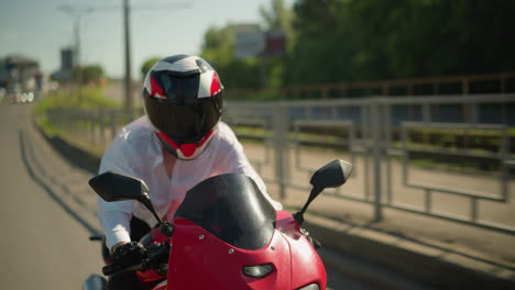 una motociclista femenina con casco monta una bicicleta eléctrica roja rápido cerca de un sendero de paseo con un carril de hierro, con un árbol verde exuberante borroso a la vista por el lado