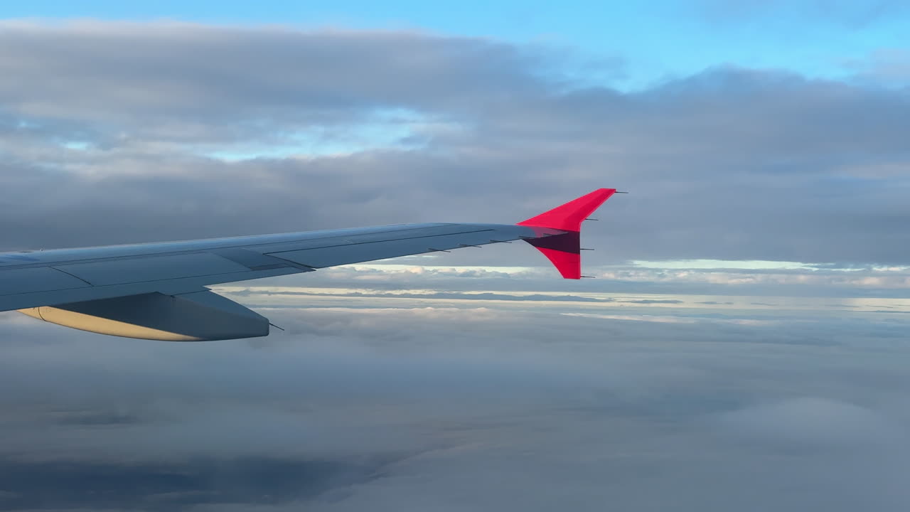 Gros plan sur l'aile d'un avion commercial traversant des nuages stratifiés, baignée d'une douce lumière naturelle. Une scène atmosphérique paisible, vue de la perspective d'un passager, bien au-dessus des nuages.