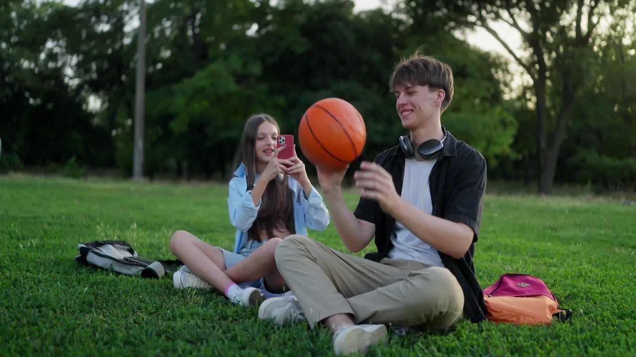 Two friends sitting on the grass in the park. One is holding a basketball and the other is taking a picture with a phone.