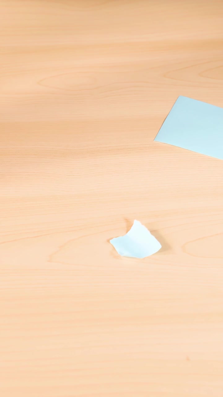 Hands remove blue origami paper scraps from a light wooden table, top-down view, bright lighting