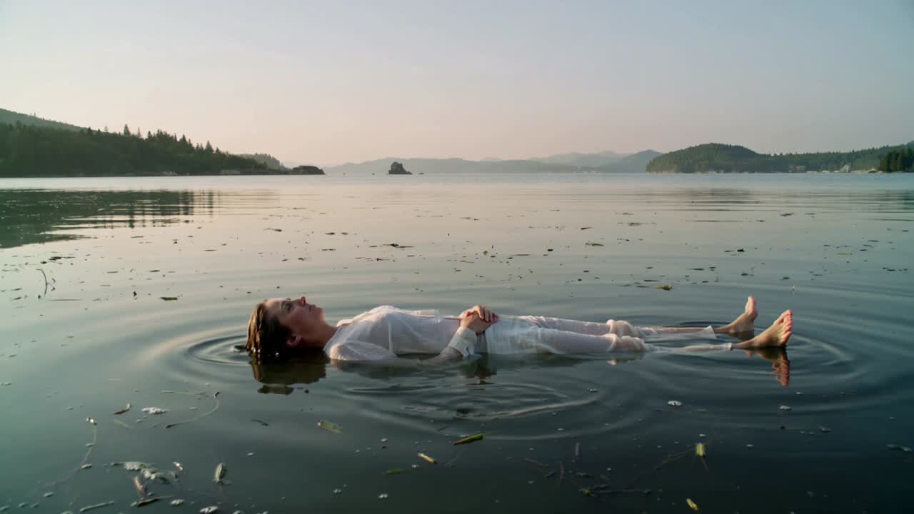 Floating adult woman adjusting legs slightly, causing ripples across calm bay, wearing white outfit
