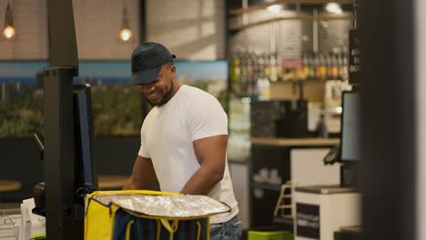 A-delivery-man-with-Black-skin-in-a-white-T-shirt-happy-and-joyful-puts-all-the-necessary-purchased-products-into-a-yellow-bag-while-standing-at-the-checkout.-Happy-male-delivery-man-buys-the-necessary-goods-and-puts-them-in-his-bag-to-deliver-to-the-client