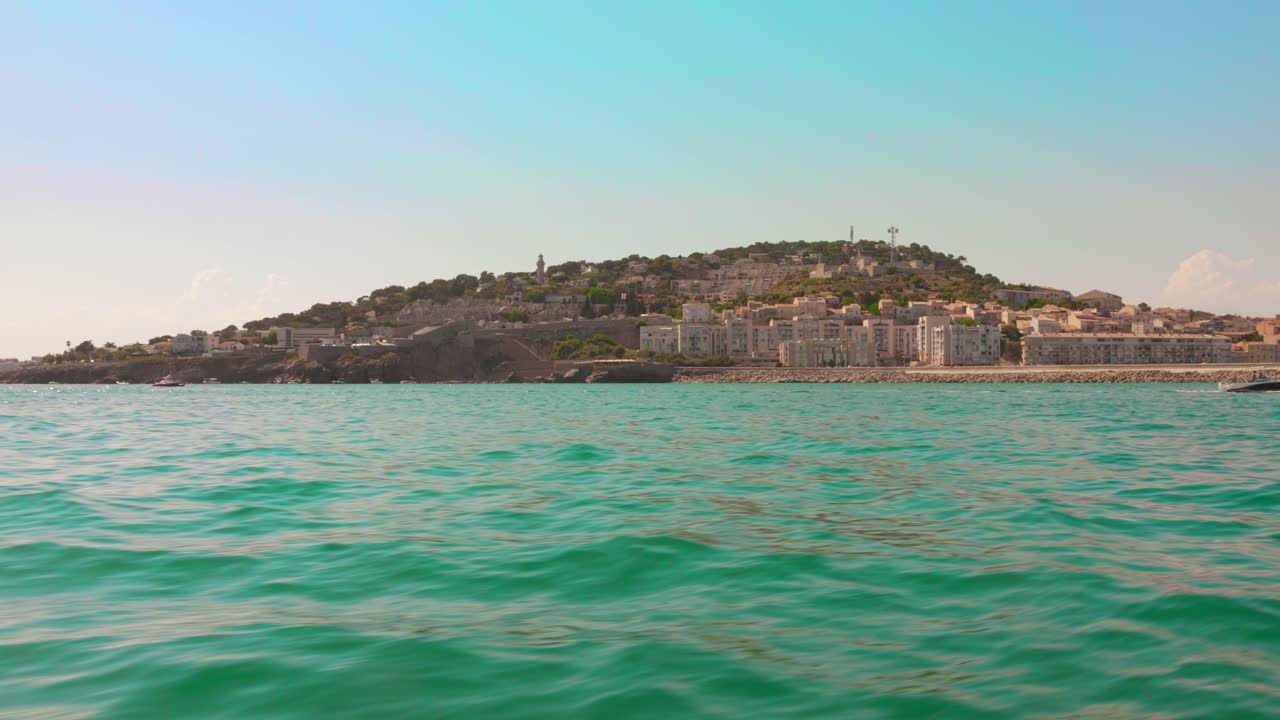 View from boat cruising along the coast of Sète in southern France, with hillside houses and turquoise Mediterranean waters under a clear sky