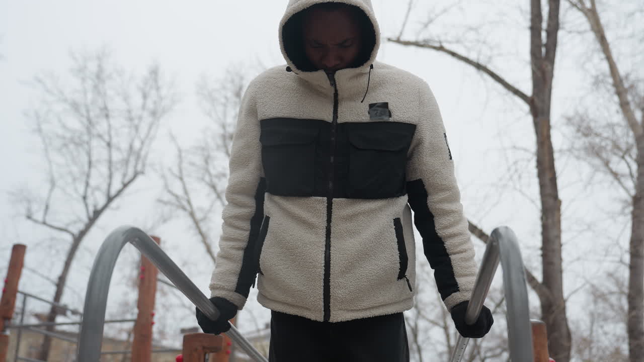 젊은이가 겨울 동안 야외 평행 바에서 근육을 쌓기 위해 푸시업 운동을 수행하는 클로즈업, 그는 후드 재과 장갑을 입고 있으며, 차가운 공기에서 눈에 띄는 숨을 쉬고 있습니다.