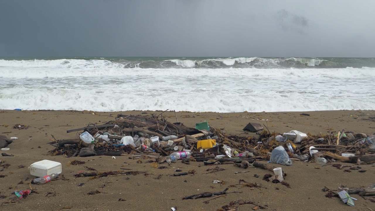 Mixed plastic containers bottles foam packaging bags and driftwood form a concentrated pile on open sand while broad white storm waves break across the entire ocean background