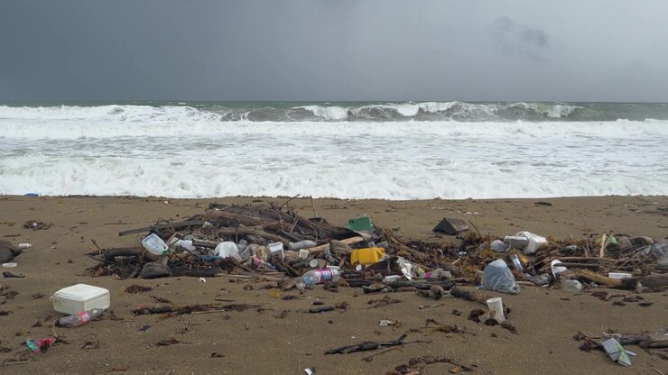 Mixed plastic containers bottles foam packaging bags and driftwood form a concentrated pile on open sand while broad white storm waves break across the entire ocean background