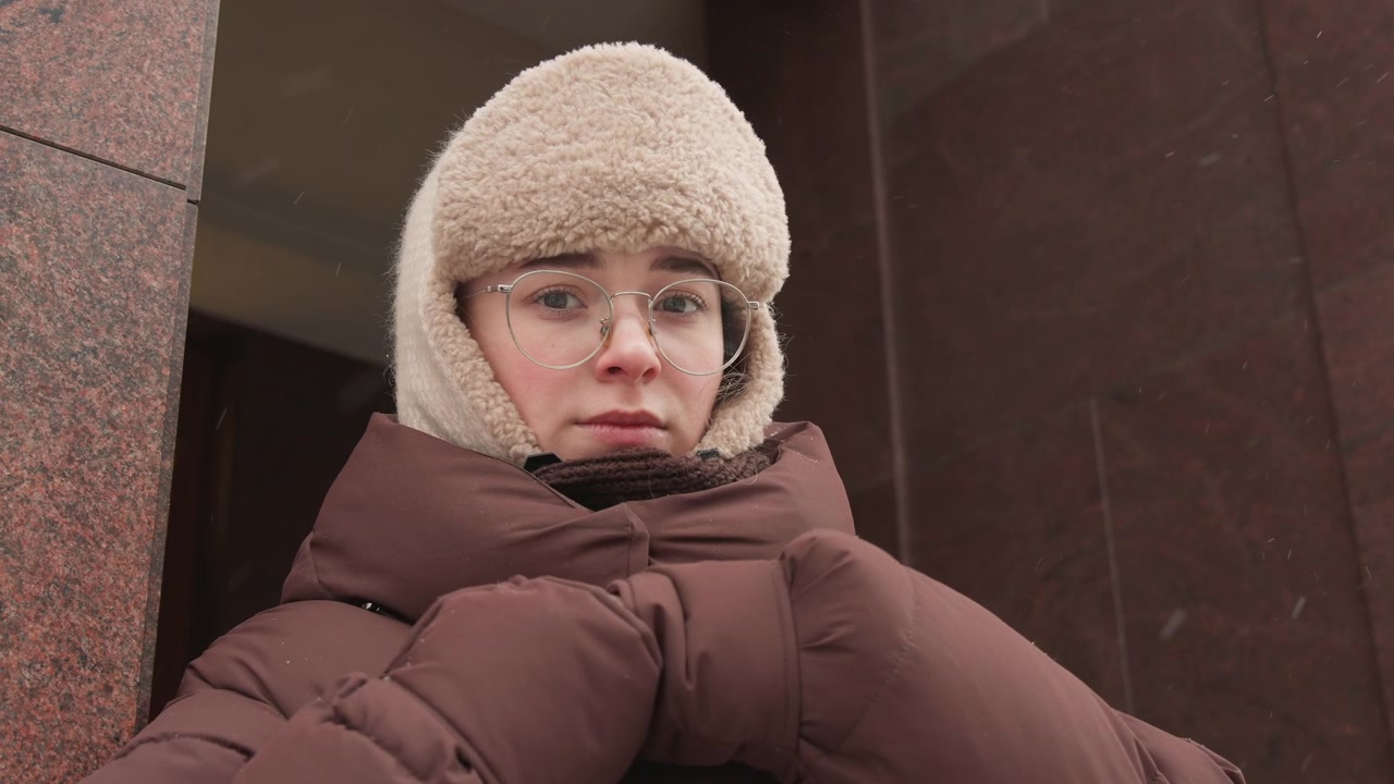 Medium close-up from low angle shows young woman wearing fluffy trapper hat and glasses, pulling insulated puffer coat collar toward chin while looking downward on overcast urban street