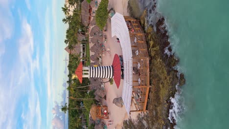 vertical drone shot of lighthouse of playa dominicus in bayahibe. orbiting shot. crashing waves of caribbean sea. people eating in restaurant. dominican republic.