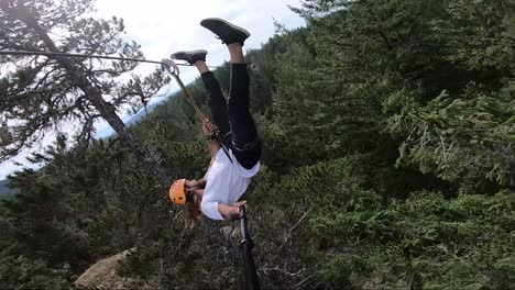 vertical action camera shot of a young man hanging upside down in a zipline in canada with big trees around and under him