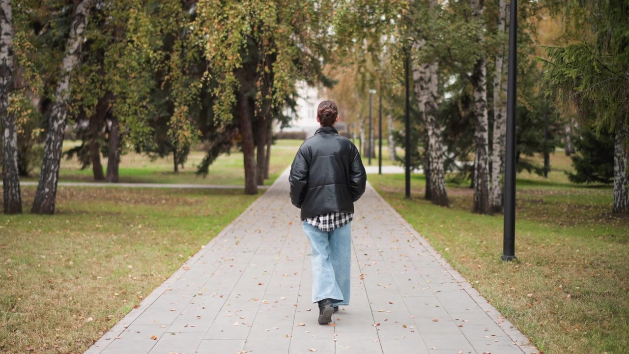 Mulher caminha firmemente por um caminho pavimentado em um parque ao ar livre, vestindo jeans azul claro de perna larga e jaqueta de couro preta. A cena apresenta árvores altas com folhagem verde em ambos os lados do caminho