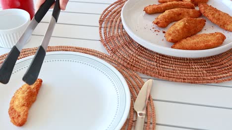 tongs place crispy fried chicken pieces onto a plate on a white table with woven placemats