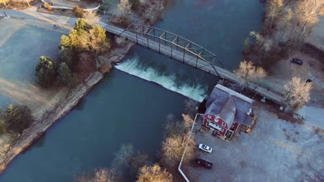 Luftaufnahme-Der-Funktionierenden-Schrotmühle-Mit-Schaufelrad,-Das-Von-Einem-Fluss-Gedreht-Wird,-Der-Von-Einer-Alten-Eisenbrücke-überspannt-Wird