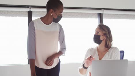 two diverse female colleagues wearing face masks looking at laptop and discussing in office