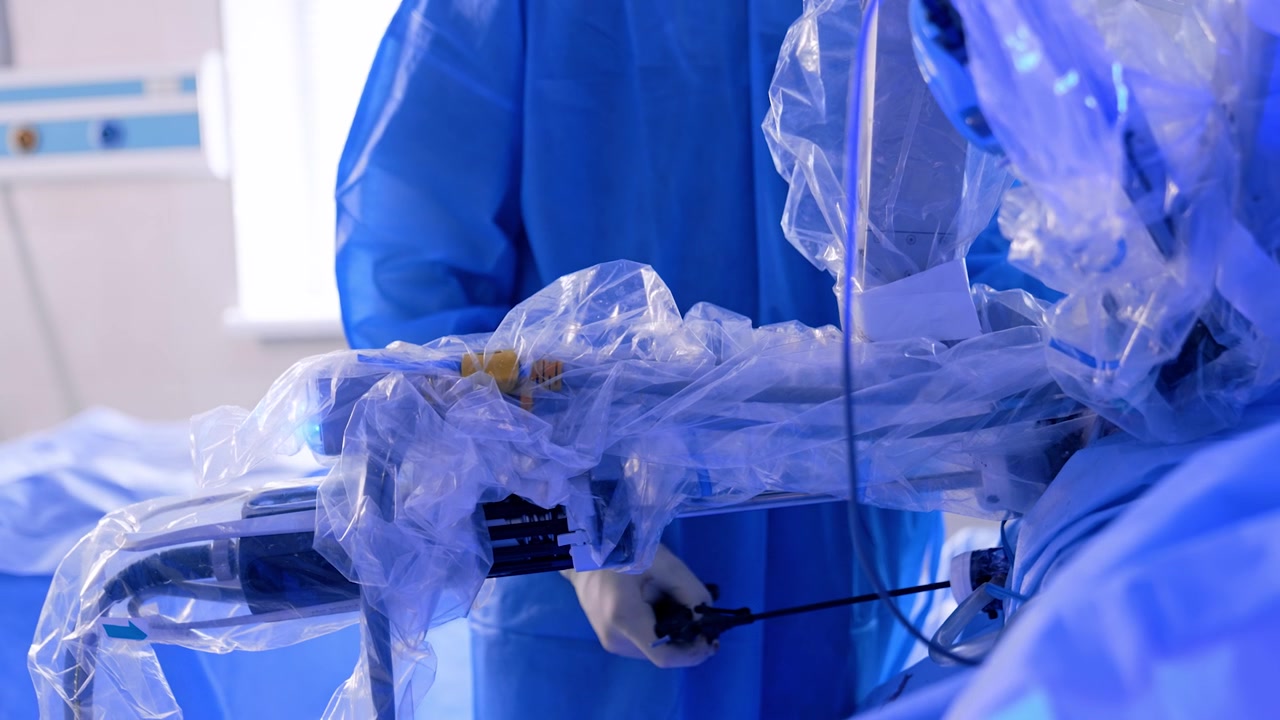 Da Vinci robot manipulator arms covered in plastic moving down at operation. Unrecognized surgeon in blue uniform stands holding the laparoscopic tool.