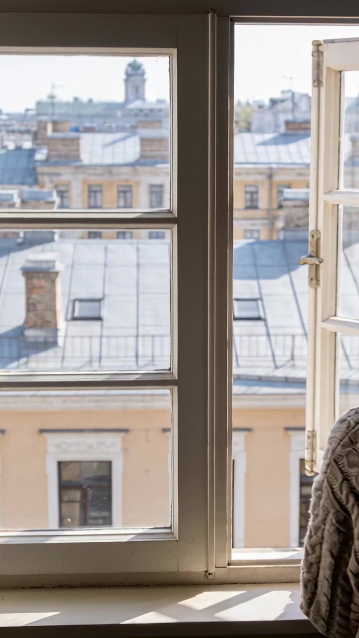Vertical video: Entering person from right in sweater standing at flat window viewing roofs for air