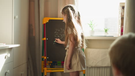 Girl-writes-math-problems-on-blackboard-teaching-brother