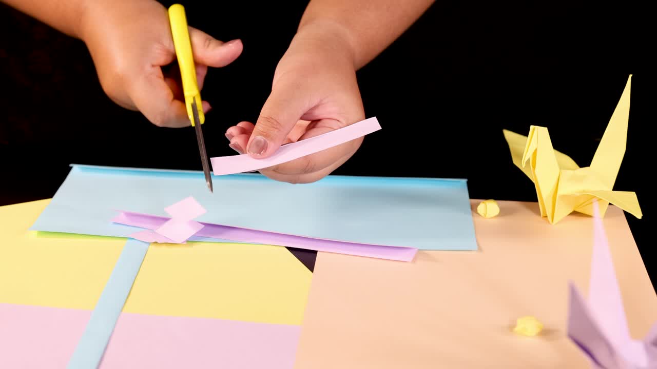 Person cuts pastel paper strips with scissors on desk, origami cranes visible, bright lighting