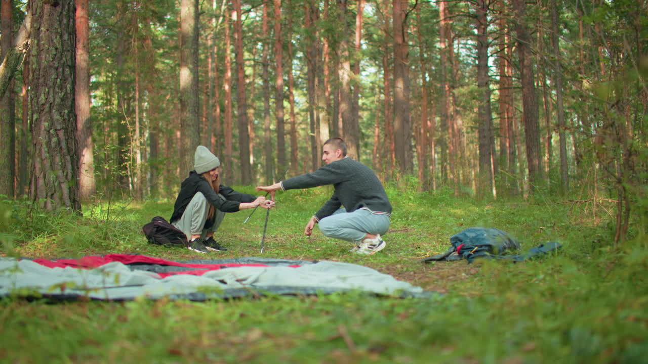 Wife playfully holding tent pole as husband collects it with smile during outdoor forest camping, surrounded by gear and nature, sharing lighthearted moment while setting up campsite together