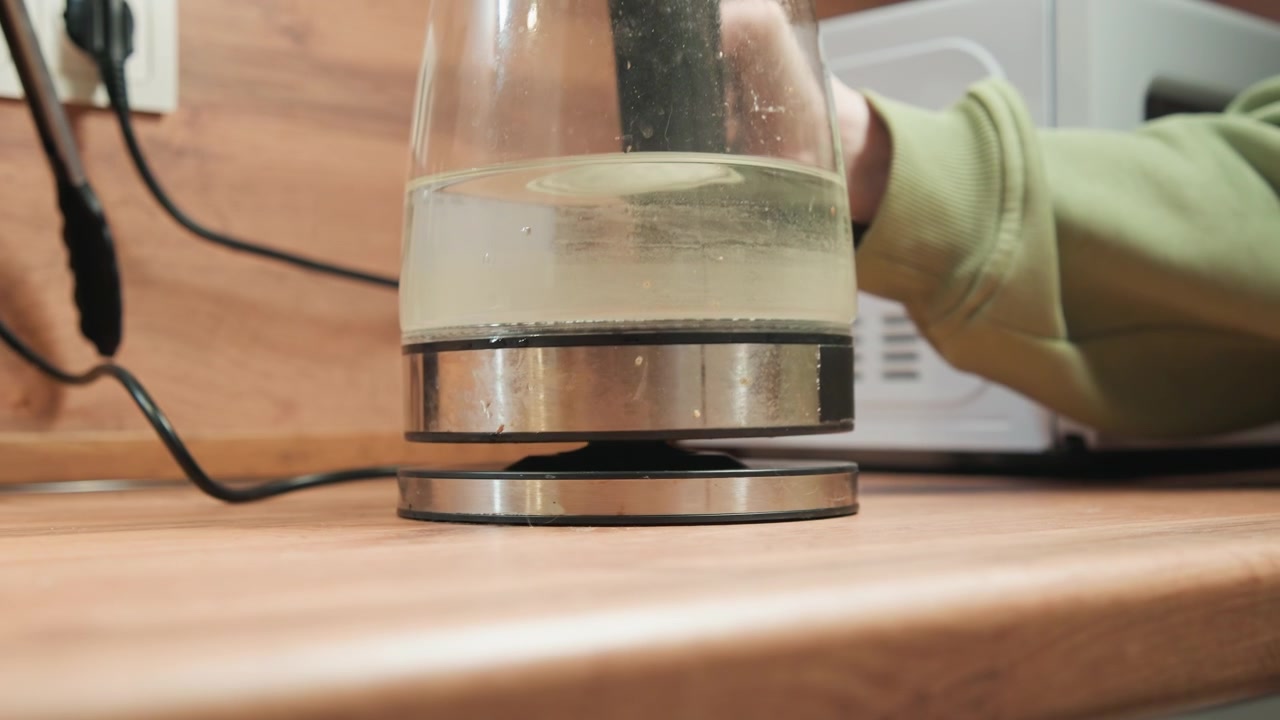Tampilan dekat sudut rendah menunjukkan ketel kaca diletakkan di atas alas pemanas logam di meja dapur kayu, dengan tangan memandu gerakan terkontrol, sorotan reflektif, dan pemisahan singkat