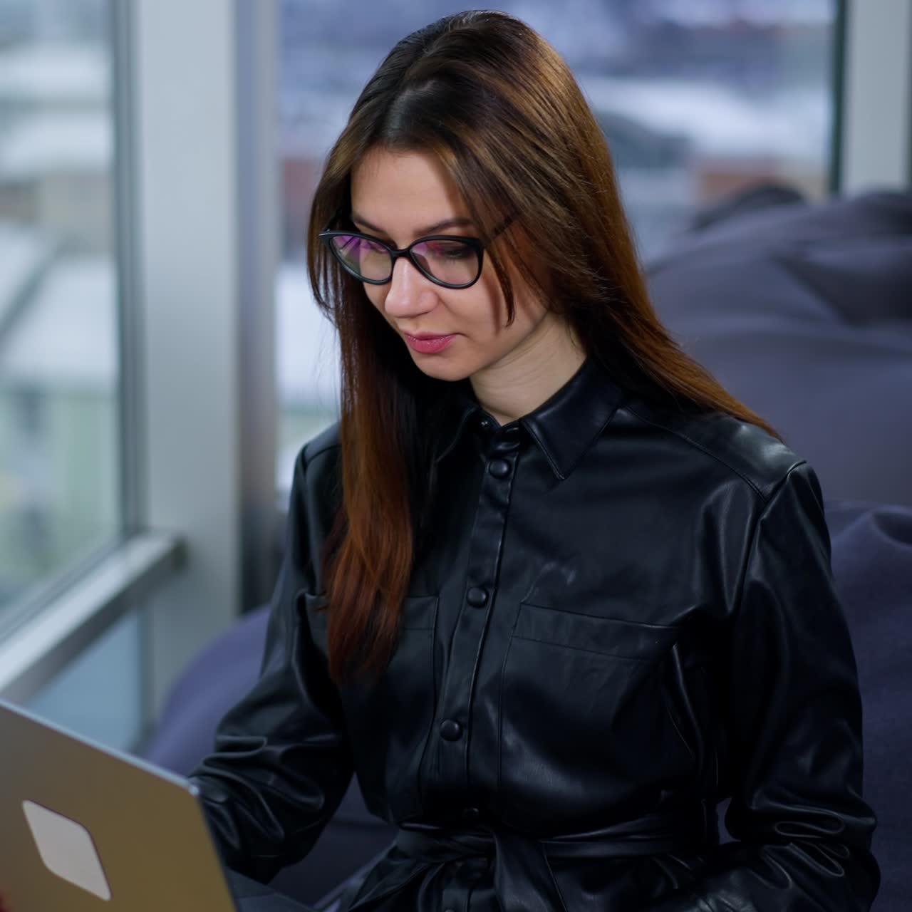 Happy smiling brunette lady sitting with laptop on her laps. Mid aged business lady in glasses working in the office at the window