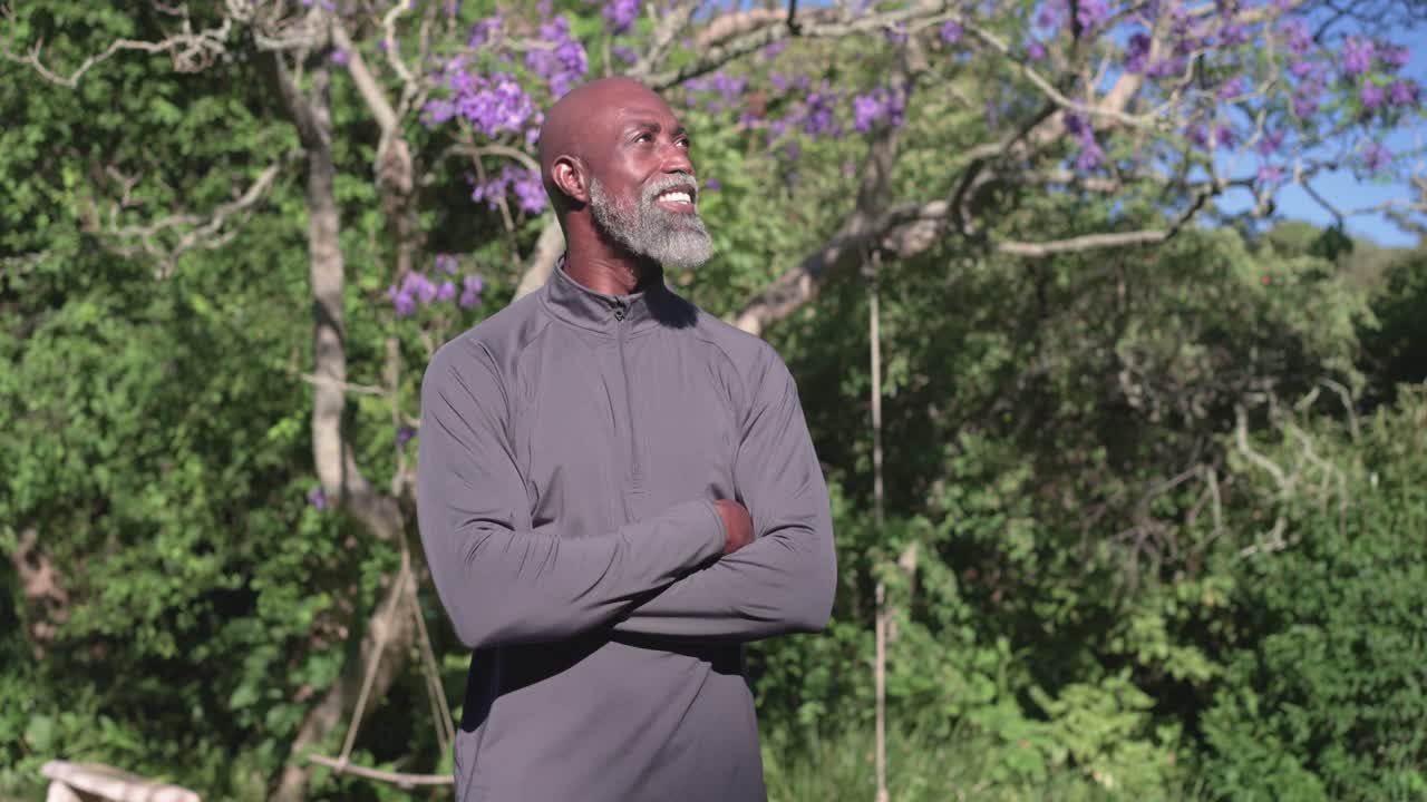African American man folding arms turning head at park under purple tree, camera zoom causing smile