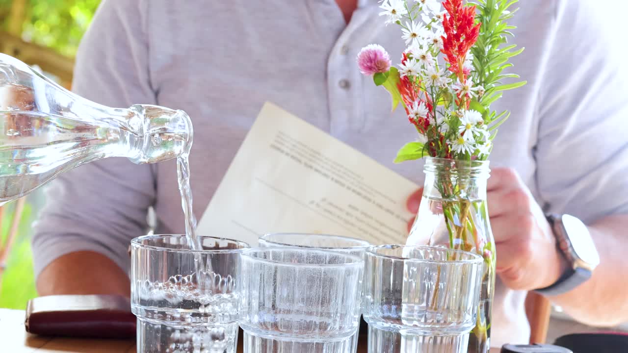 A man in a light shirt pours water from a glass pitcher into a tumbler at a sunlit outdoor café table, with flowers and menus visible