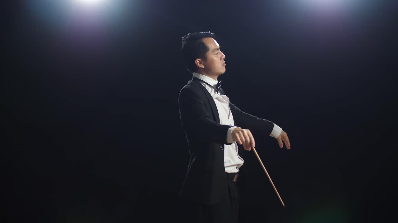 Side View Of Asian Conductor Man Holding A Baton Closing His Eyes And Showing Gesture In The Black Studio