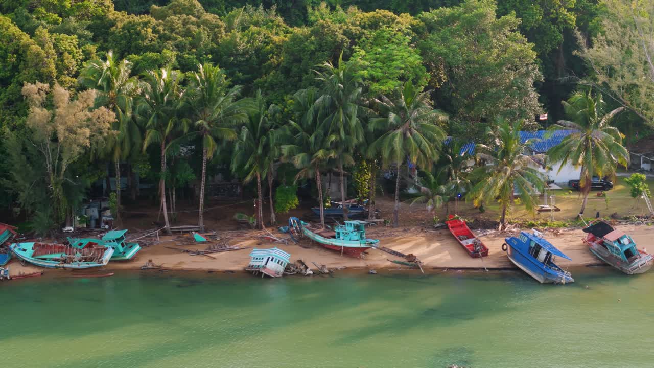 Sideways trucking drone movement along tropical shoreline with derelict fishing boats resting on sand beside palm trees and lush jungle