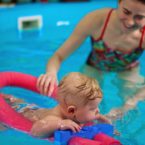 little adorable baby boy holding for swimming foam stick in the pool. woman supporting the kid learning to float in water.