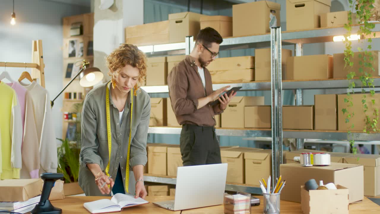 кавказская молодая женщина-дизайнер печатает на ноутбуке и пишет детали заказа на складе магазина одежды, в то время как мужчина набирает текст на планшете и проверяет коробки