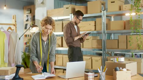 caucasian young woman designer typing on laptop and writing order details in clothing shop warehouse while male coworker typing on tablet and checking boxes