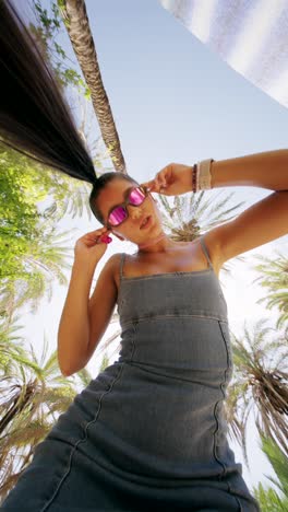 young woman with ponytail and pink sunglasses posing under palm trees