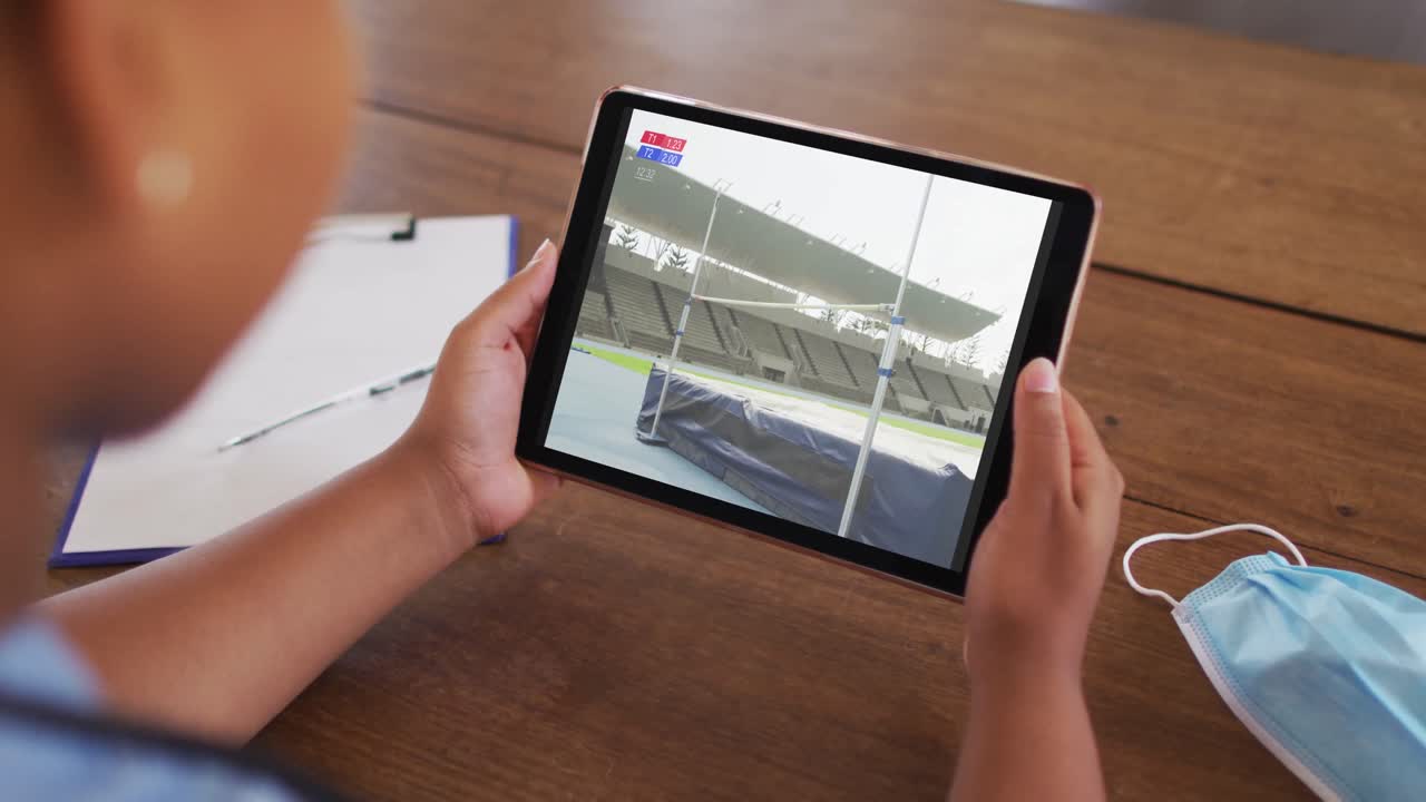 compuesto de una mujer sentada en una mesa con una máscara facial, viendo el evento de salto alto de atletismo en la tableta