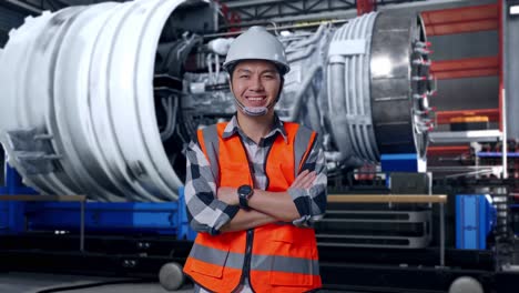 asian male engineer with safety helmet crossing his arms and smiling to camera while standing with airplane engine maintenance conducted, analytics checking the turbine