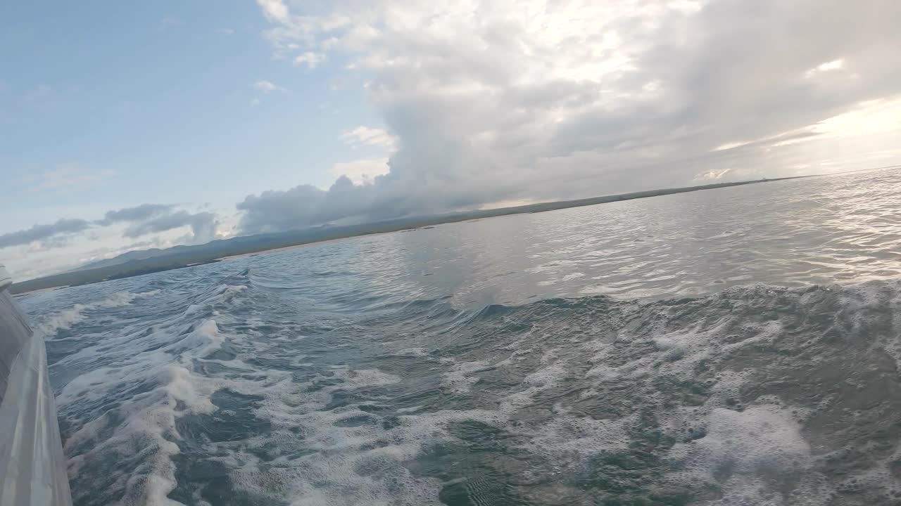 From a boat ride, an island is seen on the back. Waves are formed by the motion of the panga. Clouds and a blue sky are on the back