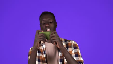 A-serious-young-african-american-man-is-playing-games-on-his-smartphone-and-doing-a-winner-gesture-standing-isolated-over-blue-wall-background-in-studio