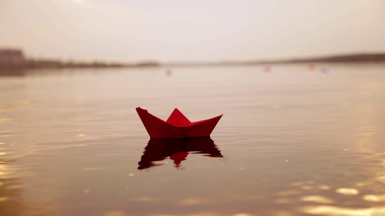 Red paper boat floating alone with the slow motion in the river at sunset. Lovely kid's origami ship on water surface in the evening. Close-up
