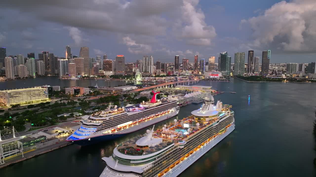 Cruise ships docked at Port of Miami with downtown Miami skyline glowing at dusk. The waterway, large vessels, and city skyline create a vibrant harbor scene. Aerial.