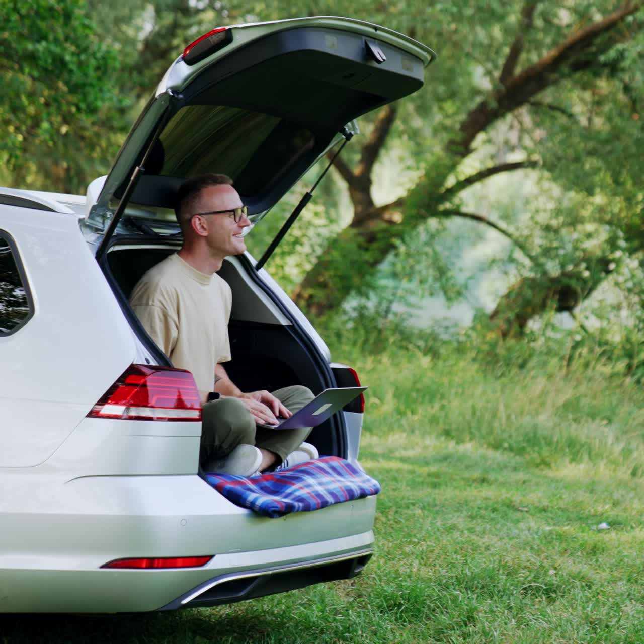 Happy man sits in his car trunk in the forest. Smiling freelancer works on laptop enjoying nature