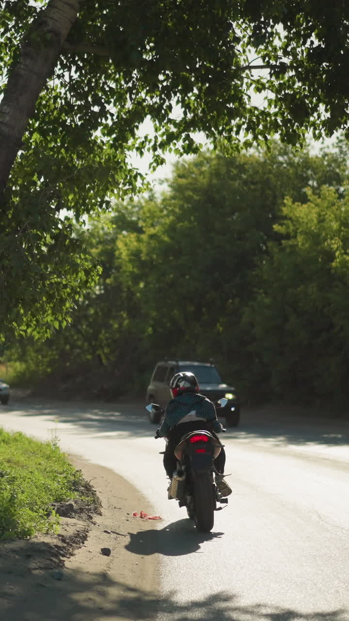 vista trasera de una motociclista femenina, su ropa revoloteando en el viento mientras monta su bicicleta. se ven coches viniendo en la dirección opuesta, con vista a coches estacionados y edificio