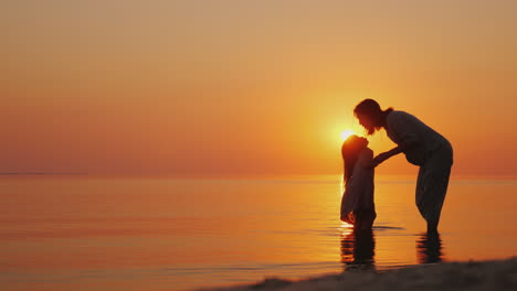 A-Pregnant-Woman-Gently-Kisses-Her-Eldest-Daughter-Beautiful-Silhouettes-Near-The-Sea-At-Sunset-Wait