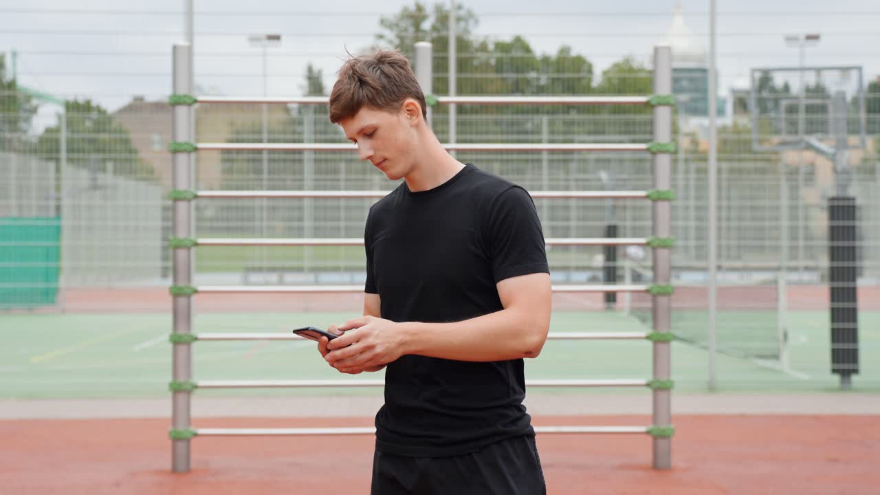 A man in black sportswear stands on an outdoor sports court, scrolling on his smartphone. The camera is static, capturing him in profile view