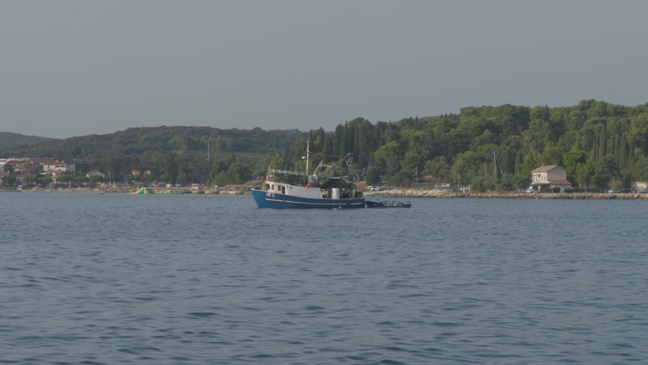 Ampia inquadratura statica di un peschereccio al lavoro al largo di Rovigno, Croazia. Acqua calma dell'Adriatico e costa verde sotto un cielo estivo luminoso