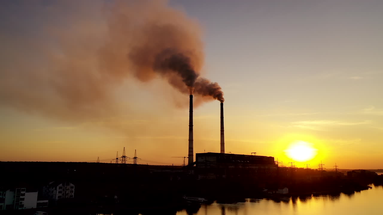 Dark power plant at the bank of the evening river. Industrial pipes of manufacturing with dark smoke at sunset. Aerial view.