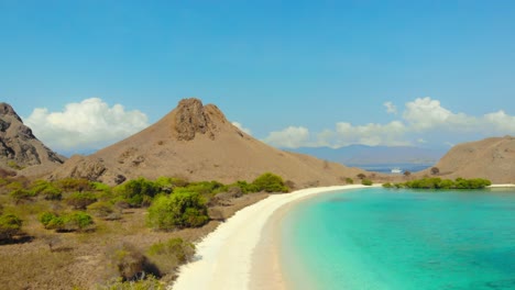 Flight-over-the-pink-beach,-Pantai-Merah,-in-Indonesia,-capturing-breathtaking-aerial-perspective-of-its-unique-and-vibrant-beauty-of-steep-mountains-and-turquoise-water-with-blue-sky-in-background