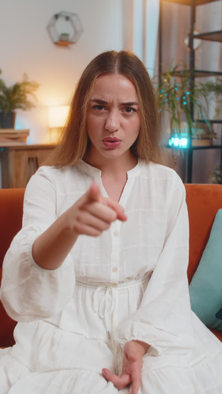 Furious young caucasian woman sitting on sofa at home angrily pointing at you through the camera