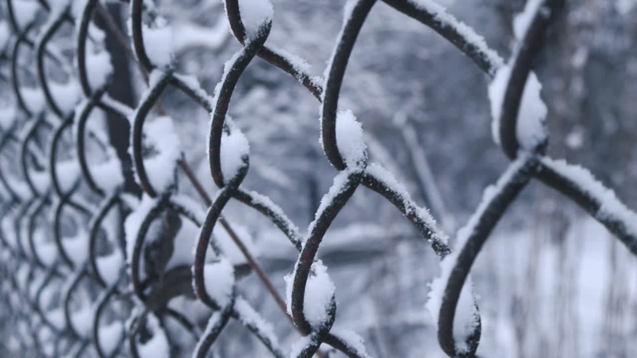 Close up footage gliding over iron metal chain link fence that has white fluffy and thick snow covering it during winter time cloudy day light. White snow covered garden in the background with foliage