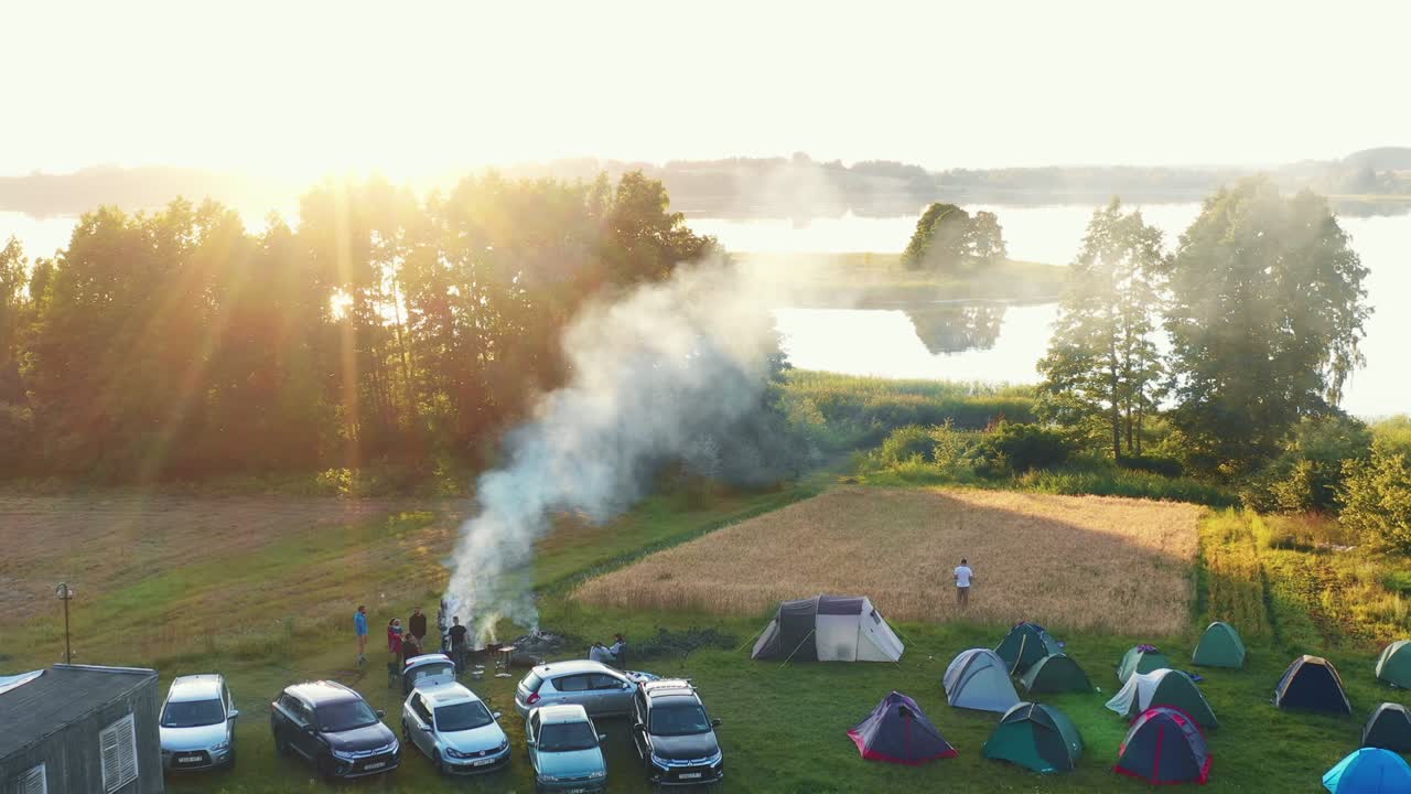 불, 자동차 및 텐트와 함께 캠핑 장소 위를 날아다니는 드론은 큰 호수와 서사시적인 여름 해가 뜨는 렌즈 플레어 파노라마를 향합니다.