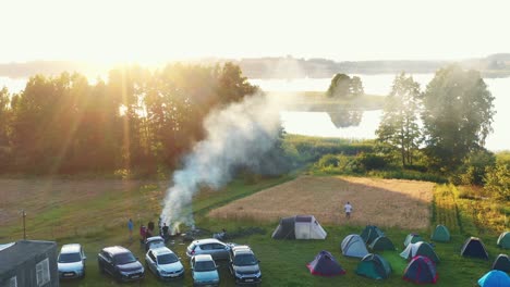 drone flying above camping spot with fire, cars and tents towards large lake and epic summer sunrise lens flare panorama